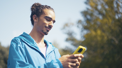 Person in bright blue jacket looking down at yellow smartphone outdoors on sunny day with trees in background