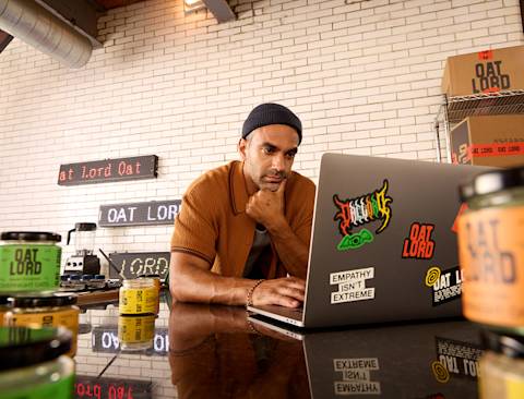 Person in brown sweater and beanie working on laptop decorated with stickers at desk with Oat Lord branded products and signage