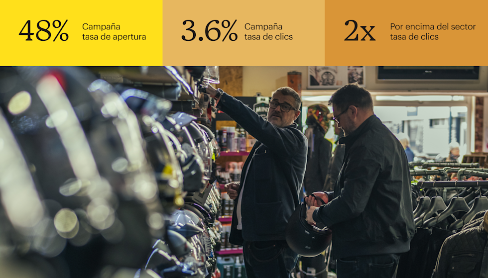 Dos hombres miran cascos en una tienda de motocicletas, ambos con chamarras oscuras y anteojos. Uno de ellos estira el brazo hacia un estante en el que se exhiben diversos cascos. El otro sostiene un casco negro.