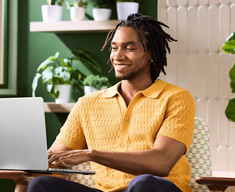 Person wearing yellow knit polo shirt smiles while working on laptop in bright, plant-filled room